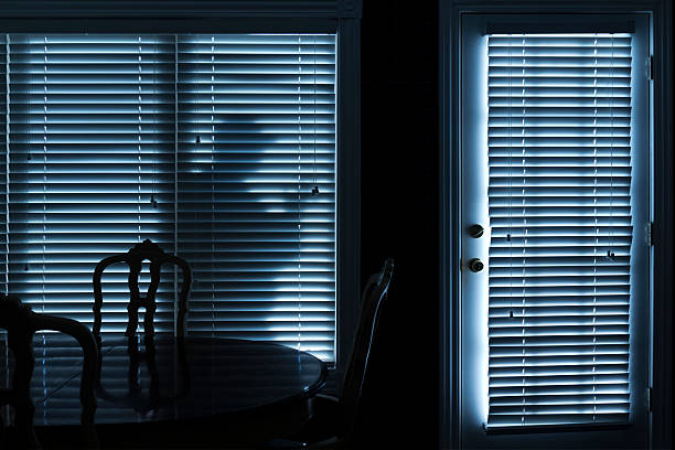 The shadow of a burglar stands on the other side of a window with blinds.